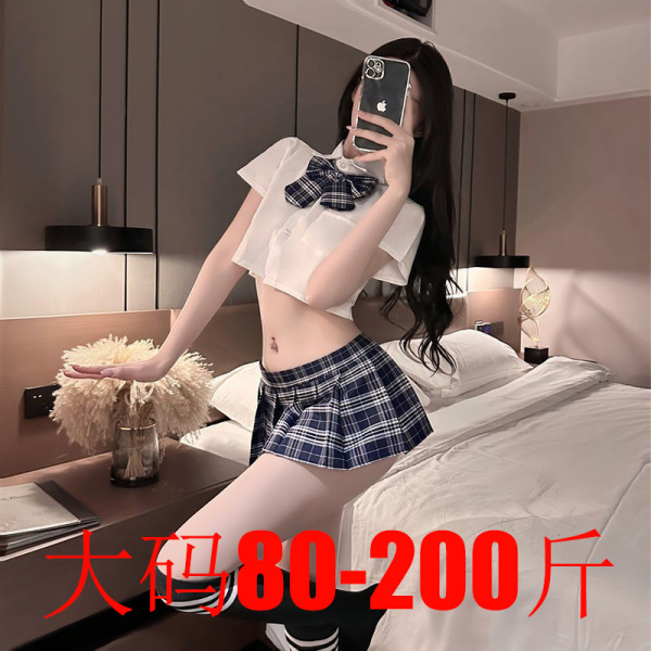 Woman modeling plus size jk uniform set with white crop top, plaid bow tie, and pleated mini skirt in bedroom.
