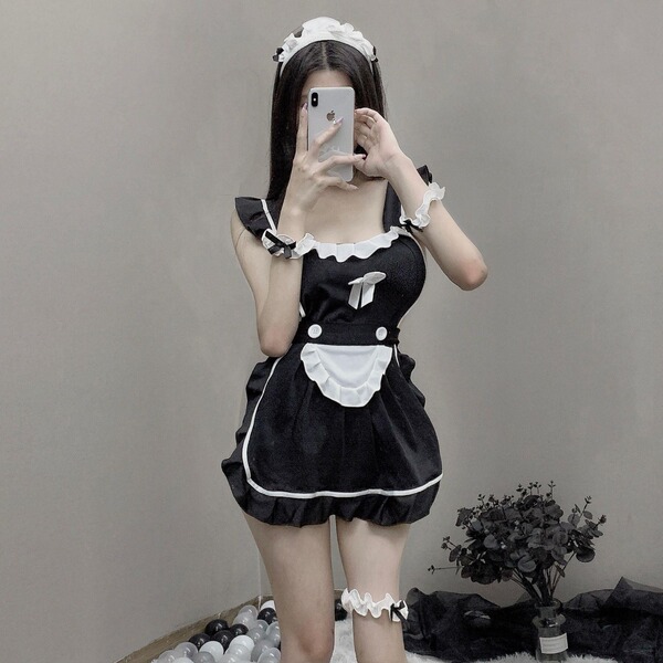 A woman poses in a black maid costume featuring lace apron, ties, and ruffled details, capturing her reflection on a smartpho...