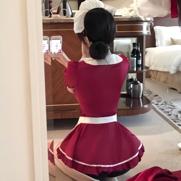 A woman wearing a red maid costume with a white apron, captured from behind as she takes a photo in a room setting.