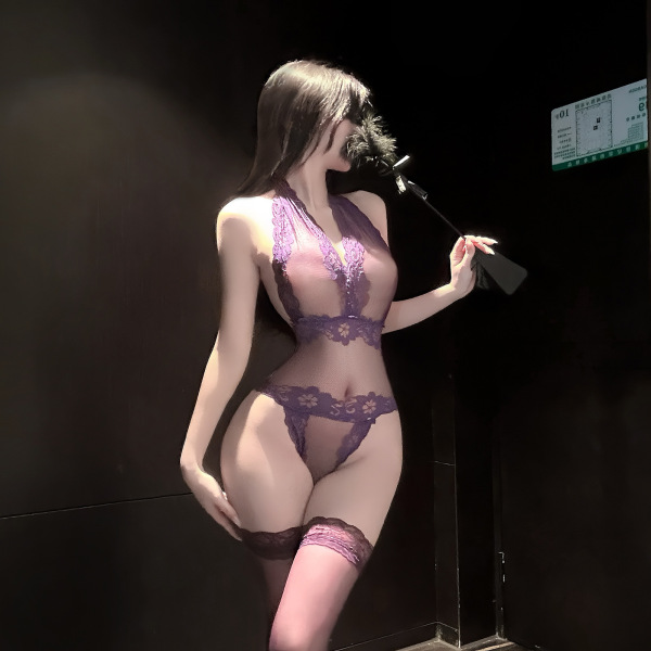 Woman modeling purple lace lingerie set with sheer top, thong, and thigh-high stockings in dimly lit room.