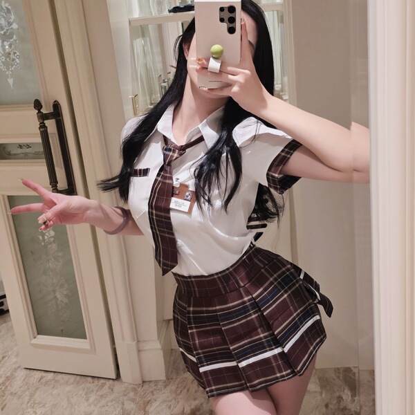 A woman poses in a mirror wearing a cosplay student uniform outfit, featuring a white blouse with a tie and a plaid skirt, em...