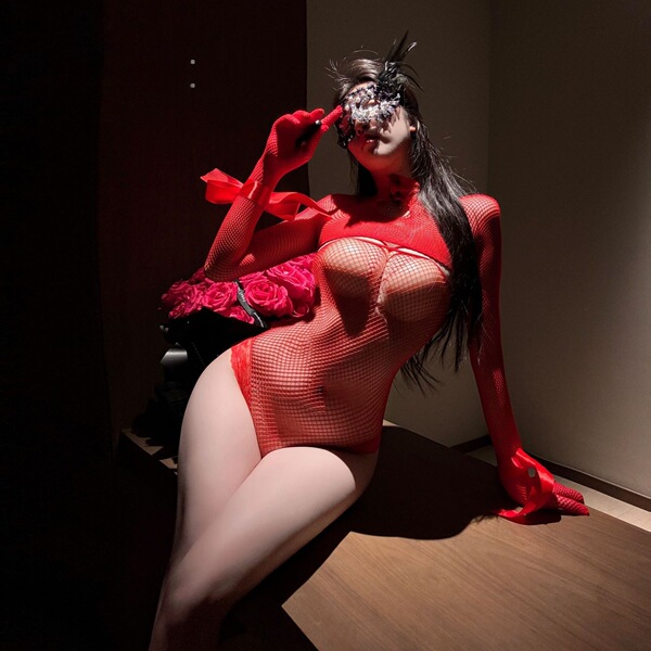 Woman wearing red sheer mesh bodysuit with long sleeves and gloves, posing seductively in dim lighting.