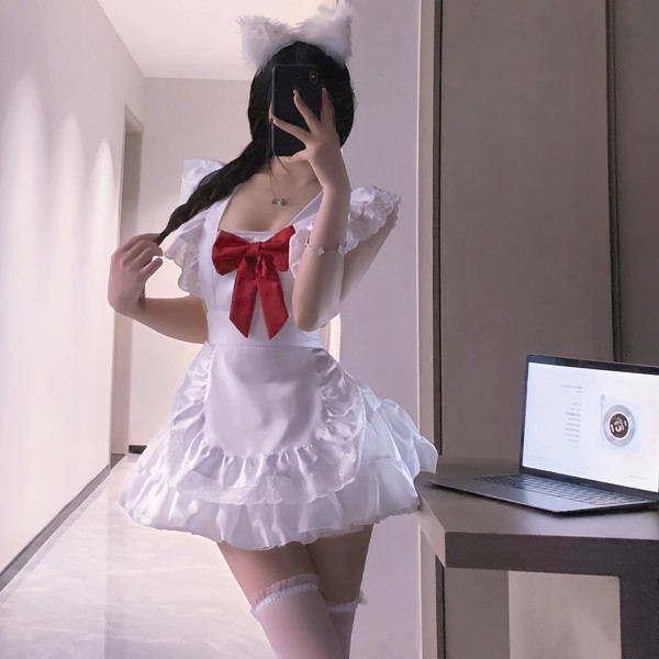 A person wearing a white maid costume with a red bow, fluffy cat ears, and thigh-high stockings poses indoors next to an open...