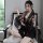 A woman sits elegantly on a chair wearing a black floral-patterned kimono-style robe, paired with a matching obi belt. the ou...
