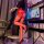 Woman wearing sheer red open-crotch bodystocking with black heels, posing by gaming chair and monitor in dimly lit room.