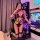 Woman in black leather maid costume with bondage straps, fishnets, and choker, posing in gaming room with rgb lights.