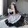 A person wearing a black and white japanese maid costume, featuring ruffled sleeves and skirt, poses with a phone against a b...