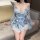 Woman in light blue floral seductive maid costume lingerie set with sheer robe and bow, sitting on sofa holding fan.