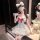 A woman poses in a maid-inspired lolita outfit featuring a white dress with lace details, a red bow, and cat ears. she wears ...