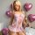 A woman poses in a sheer pink lace bodysuit adorned with bows and cut-outs, holding a rose against a backdrop of heart-shaped...
