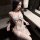 Woman wearing midnight desire lace bodysuit, sheer white lingerie with ruffles and open-crotch design, seated in dim room.