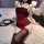 Woman wearing red and black lace bodysuit with garter straps, seated on bed with rose petals and whip nearby.
