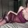Woman wearing purple lace bodysuit and matching sheer skirt, lying on bed in elegant lingerie set.