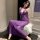 Woman wearing purple sheer bodysuit with lace trim and bow details, seated on white fur stool in elegant room setting.