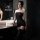 Woman wearing black sheer bodysuit with lace trim and garter straps in modern bathroom setting.