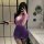 A woman poses in a bathroom wearing a purple ol secretary lingerie set, featuring a deep v neckline, sheer lace bodice, and a...