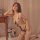 Woman wearing qingyege sheer mesh bodysuit with suspender stockings, posing beside a wooden table with pink flowers.