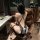 Woman in sheer black bunny girl bodysuit with straps and open-crotch design, seated on leather chair.