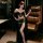 Woman modeling luxury black lingerie set with fringe bra, sheer robe, and gold chain belt in elegant room.